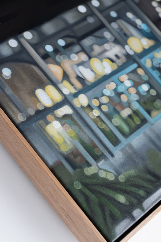 A close-up angled view of a framed art print or decorative panel with a light wood frame and a glass surface showing an abstract cityscape or window-like scene in muted blue, green, yellow, and gray tones.