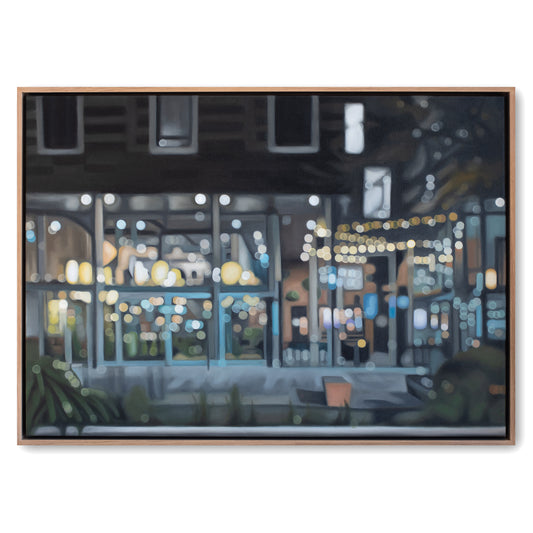 A framed painting depicting a softly blurred nighttime street or restaurant scene with glowing windows, warm string lights, and cool blue-gray tones, shown against a plain white background.