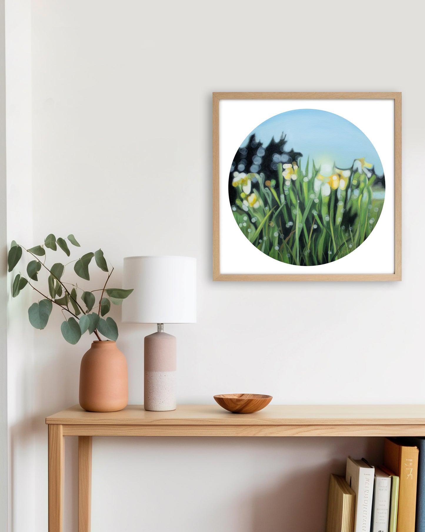 Spring daffodil flowers in a blurry bokeh style framed in a wooden frame hanging above a wood bookshelf, this fine art print is fresh and yellow with green. Painted in a blurry bokeh style by New Zealand bokeh artist Mel McKenzie, The Penny Drops.