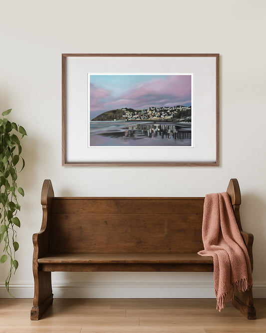 Wooden bench with a pink blanket against a wall with a framed picture of a coastal scene - St Clair Beach, Dunedin by New Zealand artist, Mel McKenzie of The Penny Drops.