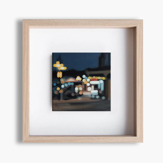 Wanaka New Zealand, lakefront drive cafe. Painted in a blurry bokeh style on a small canvas framed in wood frame. Lights of the street and stores. Art by New Zealand artist Mel McKenzie, The Penny Drops.