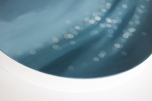 Art on round canvas framed in white, water bokeh, shimmering light on ocean or lake. Blue painting by Mel McKenzie, The Penny Drops.