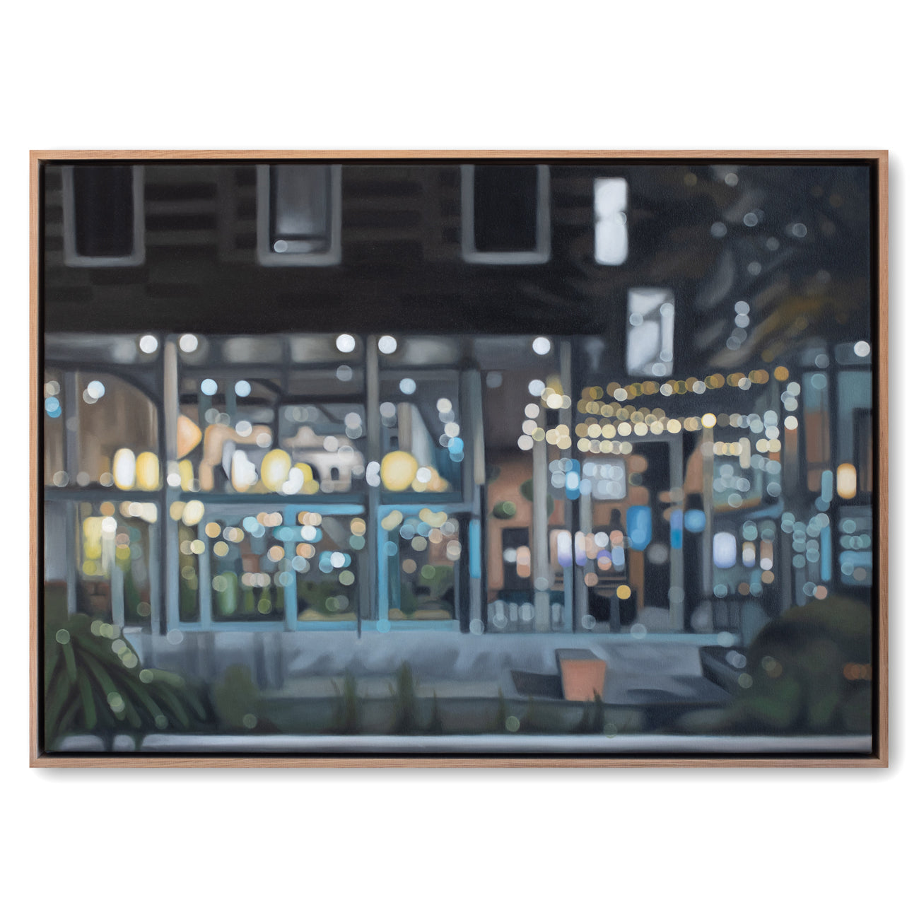 A framed painting depicting a softly blurred nighttime street or restaurant scene with glowing windows, warm string lights, and cool blue-gray tones, shown against a plain white background.