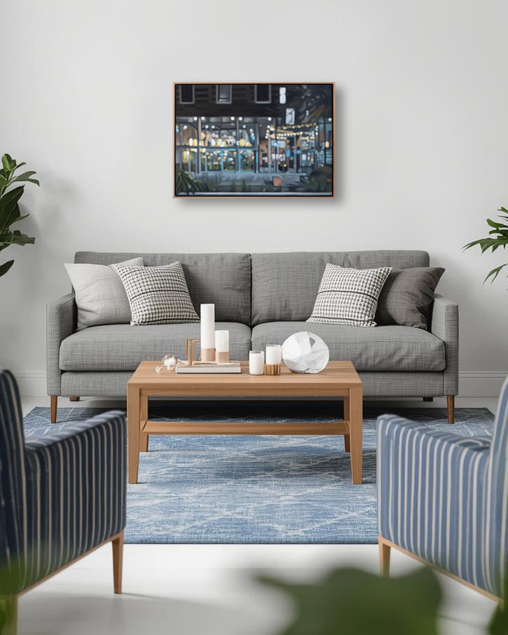 A framed horizontal canvas print of a city street at night with warm lights, displayed on a white wall above a gray sofa in a modern living room setting.