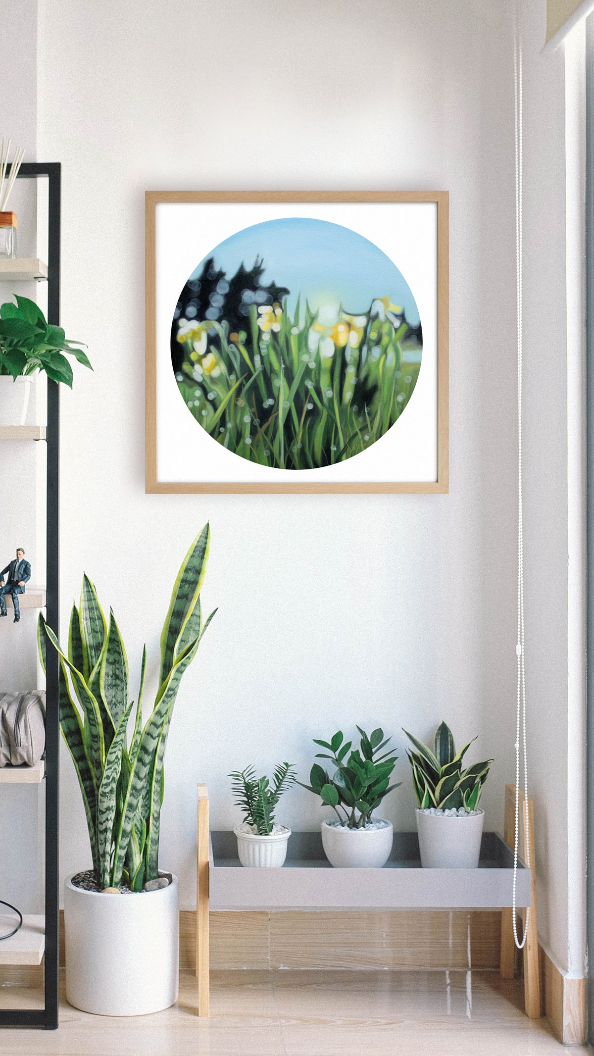 Spring daffodil flowers in a blurry bokeh style framed in a wooden frame and hanging on a white wall above some indoor plants, this fine art print is fresh and yellow with green. Painted in a blurry bokeh style by New Zealand bokeh artist Mel McKenzie, The Penny Drops.